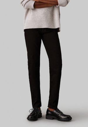 Sorte slim-fit jeans med en glat overflade, parret med sorte snøresko og en lys grå striktrøje med ribbede manchetter og kant.