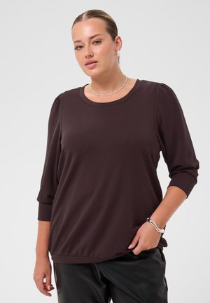 Femme aux cheveux tirés en arrière, portant un haut à manches longues marron foncé, un pantalon noir, un collier de perles et un bracelet en argent, debout devant un fond uni.