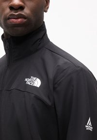 Κοντινό πλάνο ενός ατόμου που φορά μαύρο μπουφάν με φερμουάρ, με το λογότυπο The North Face στο στήθος και το λογότυπο Mountain Athletics στο μανίκι.