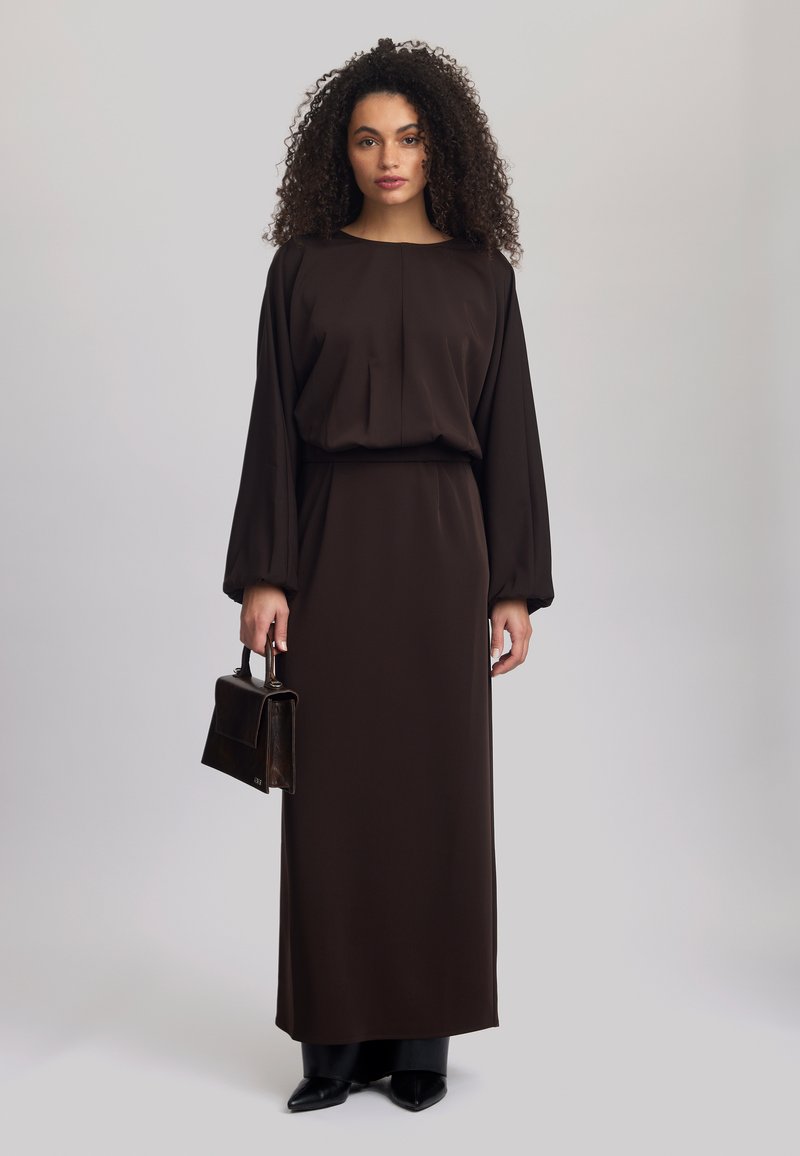 Braunes Maxikleid mit langen Ärmeln, tailliertem Schnitt, plissierten Details und glatter Textur. Kombiniert mit einer kleinen braunen Handtasche und schwarzen spitzen Schuhen.
