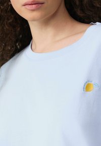 T-shirt en coton bleu clair avec un col rond, présentant un design de citron jaune brodé avec des feuilles vertes sur la poitrine gauche.