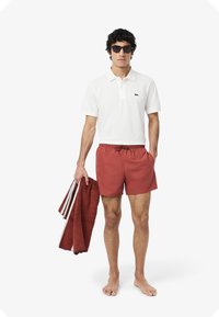 Polo bianco con un piccolo logo verde, pantaloni da bagno rossi con coulisse, mentre tiene un asciugamano rosso. Gli occhiali da sole completano il look pronto per la spiaggia.