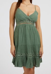 Vestido verde sin mangas con un cuerpo retorcido, acentos de encaje en la cintura y un dobladillo con volantes. Tela ligera con un acabado texturizado.