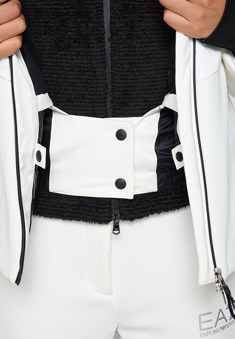 Giacca esterna bianca con zip nera, dotata di un interno in pile nero e chiusura a pressione bianca. Texture liscia con dettagli a contrasto.