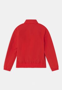 Červená větrovka s límcem, elastickým pasem a dlouhými rukávy. Hladká textura, minimalistický design, bez viditelných kovových částí nebo doplňků.