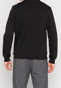 Felpa nera con maniche lunghe, colletto rotondo e polsini a coste. Texture liscia, design minimale, abbinata a pantaloni a righe grigie.
