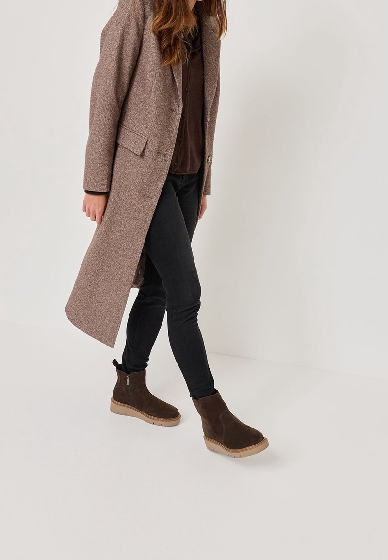 Femme portant un long manteau en laine marron, une chemise marron foncé, un jean noir ajusté et des bottines en daim marron foncé, marchant devant un fond uni.