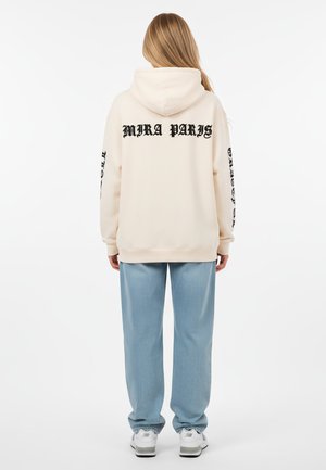 Felpa crema con cappuccio e testo nero "MIRA PARIS" sulla schiena e sulle maniche. Abbinata a pantaloni azzurri chiari e sneakers bianche.