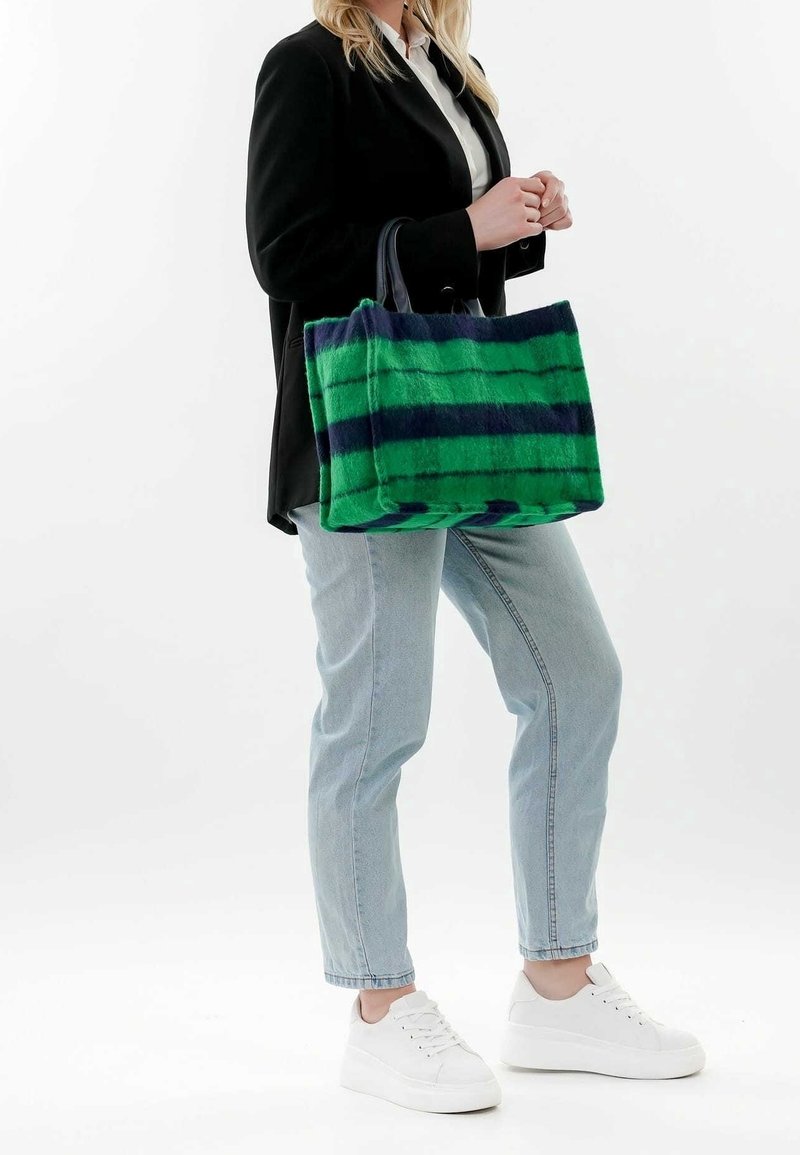 Femme portant un blazer noir, une chemise blanche, un jean bleu clair et des baskets blanches tenant un grand sac cabas rayé vert et bleu marine.