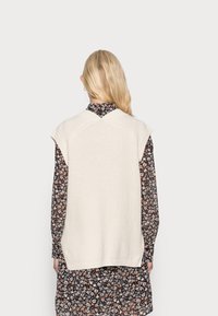 Pletený krémový svetřík bez rukávů s texturovaným vzorem a žebrovaným lemem přes hnědé šaty s květinovým vzorem a dlouhými rukávy.