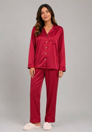 Femme debout portant un haut de pyjama en satin rouge à boutons avec manches longues et un pantalon assorti, avec des chaussons rose clair sur un fond uni.