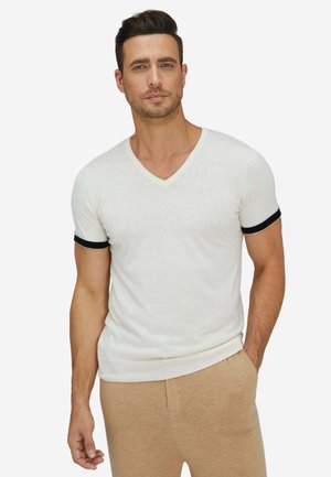 Mann trägt weißes V-Ausschnitt Kurzarmshirt mit schwarzem Saum an den Ärmeln und beigen Hosen, steht mit einer Hand in der Hosentasche.