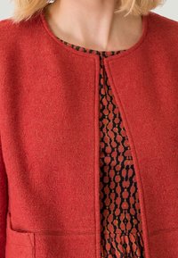 Červená texturovaná bunda s kulatým výstřihem, otevřeným předním dílem a kapsami. Pod ní černé šaty s geometrickým oranžovým vzorem.