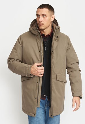 Brun, vadderad parka med huva, snap och dragkedja, samt två framfickor. Bärs över en mörk tröja och blå jeans.