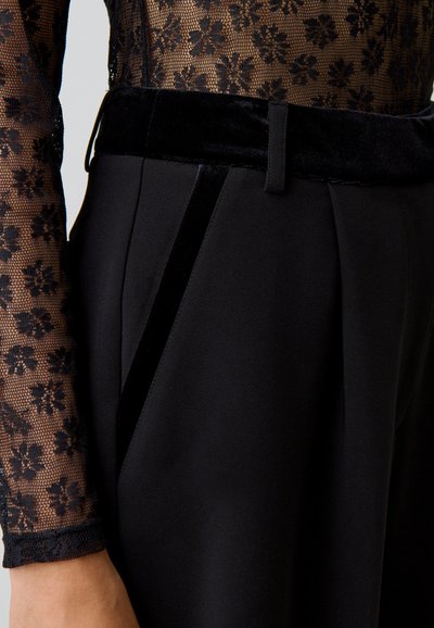 Gros plan sur un pantalon noir avec une taille en velours et un liseré sur le côté, associé à une manche en dentelle florale transparente sur un bras relevé.