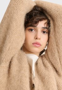 Cardigan in lana beige con una morbida texure, caratterizzato da un profondo scollo a V, grandi bottoni e una vestibilità ampia, indossato sopra una maglietta bianca.