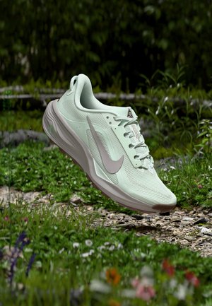 Ανοιχτό πράσινο αθλητικό παπούτσι Nike με γκρι λογότυπο, που αιωρείται πάνω από ένα μονοπάτι με βότσαλα περιτριγυρισμένο από χορτάρι και άγρια λουλούδια.