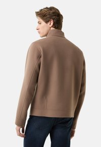 Bruine fleecejas met een hoge kraag, strakke lijnen en accenten langs de zijnaad, gecombineerd met een blauwe spijkerbroek, gezien vanaf de achterkant.