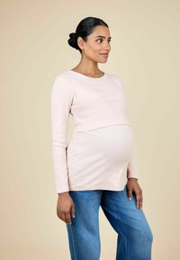 Heleda roosa pikkade varrukatega raseduse topp ribakonstruktsiooniga, komplementeeritud mugavate siniste teksadega. Mudel seisab külg ees beeži tausta vastu.