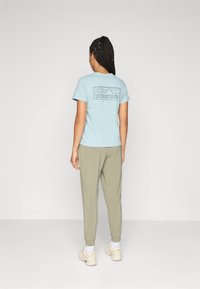 Vrouw met gevlochten haar, gekleed in een lichtblauw Patagonia T-shirt, olijfgroene broek, witte sokken en beige sneakers, staand tegen een effen achtergrond.