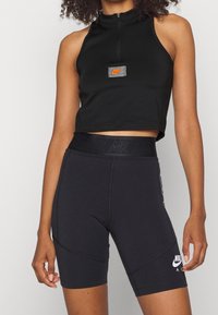 Kobieta w czarnej, bezrękawnikowej, skróconej bluzce i czarnych, wysokich szortach rowerowych Nike Air, stojąca na tle jednolitego szarego koloru.