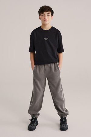 Pojke stående med händerna i fickorna, iklädd en svart T-shirt med texten "Salty Dog", grå mjukisbyxor med vita ränder och svarta sneakers mot en ljus bakgrund.