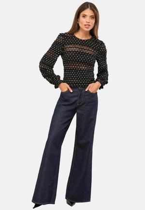 Schwarzes Top mit weißen Punkten, durchsichtigen Einsätzen und Puffärmeln, kombiniert mit dunkelblauen Jeans mit weitem Bein. Beide Kleidungsstücke haben eine lässige Passform.