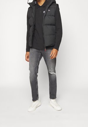 Man draagt een zwart donsvest over een zwarte poloshirt met lange mouwen, grijze gescheurde jeans en witte sneakers, staand tegen een effen witte achtergrond.