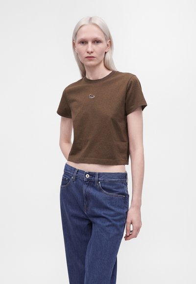 Ξανθιά γυναίκα με καφέ κοντό t-shirt με μικρό λευκό λογότυπο και ψηλόμεσα μπλε τζιν, στέκεται σε απλό λευκό φόντο.