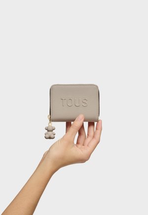 Mano sosteniendo una cartera TOUS beige con superficie texturizada y tirador de cremallera en forma de oso sobre un fondo gris liso.