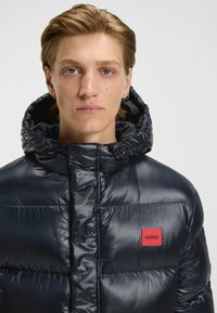 Giacca puffer di un blu scuro con finitura lucida, cappuccio regolabile e una patch logo rettangolare rossa sul lato sinistro del petto.