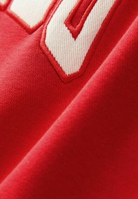 Červená tkanina s hladkým povrchem, zdobená výraznými bílými vyšívanými čísly. Design zahrnuje diagonální záhyb v materiálu.