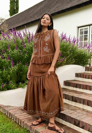 Woman in brown sleeveless top and long skirt with floral embroidery stands on brick steps near purple flowers and white building.
