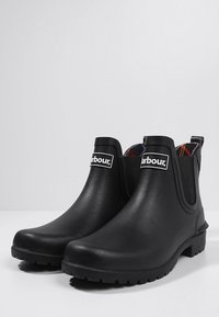 Bottes en caoutchouc noires jusqu'à la cheville avec panneaux latéraux élastiques, semelle texturée et détail de logo. L'intérieur présente un revêtement à motifs.
