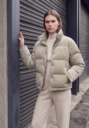 Jeune femme en doudoune beige et pantalon assorti, appuyée d’une main sur un pilier devant un rideau métallique fermé.
