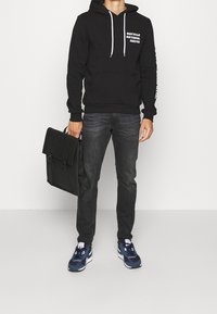 Sudadera negra con el texto "REUTILIZA, RECICLA, DEVUELVE", jeans grises, zapatillas deportivas navy y una bolsa de mensajero negra. Tejido texturizado y corte regular.