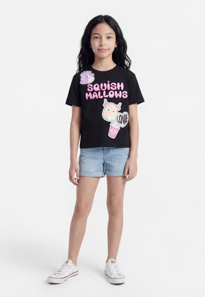 Νεαρή κοπέλα που φοράει μαύρο μπλουζάκι με ροζ γράμματα "Squish Mallows" και πολύχρωμες καρφίτσες χαρακτήρων, τζιν σορτς και λευκά αθλητικά παπούτσια.