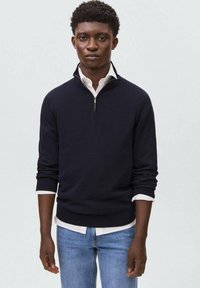 Mornarica pulover z zadrgo in visokim ovratnikom, izdelan iz mehkega blaga, s dolgimi rokavi in rebrastimi manšetami. Oblačen je čez belo srajco s ovratnikom in modre kavbojke.