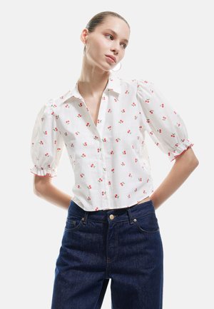 Blusa bianca con stampa di ciliegie rosse, maniche a sbuffo corte con bordo arricciato, colletto, abbinata a jeans in denim blu scuro.