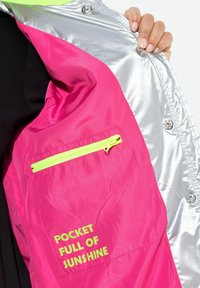 Veste coupe-vent rose et argent avec une poche zippée dotée d'une fermeture éclair vert fluo et du texte "POCKET FULL OF SUNSHINE". Texture lisse.