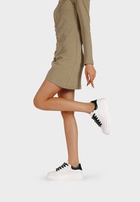 Sneakers bianchi con suola spessa, dettagli neri sul tallone e sulla parte superiore. Presentano lacci neri e una superficie testurizzata. Indossati con un abito beige.