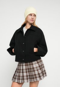 Μαύρο κοντό σακάκι με κουμπωτό κλείσιμο και μεγάλες τσέπες, συνδυασμένο με γκρι τοπ και καρό πλισέ καφέ φούστα, στολισμένο με ανοιχτό κίτρινο σκουφάκι.