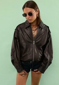 Giacca oversize in pelle marrone con zip frontale, ampio colletto e spalle strutturate, indossata sopra un top verde e pantaloni shorts in denim scuri.