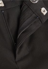 Tissu noir avec une surface texturée, doté d'une fermeture éclair à l'avant et d'une fermeture à bouton-pression. Des poches supplémentaires sont visibles, mettant en avant un design fonctionnel.