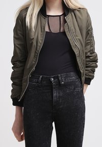 Nainen, jolla on yllään oliivivihreä bomber-takki mustan verkkopaidan päällä ja korkeavyötäröiset haalistuneet mustat farkut, seisoo rentouttavasti yksi käsi sivulla.