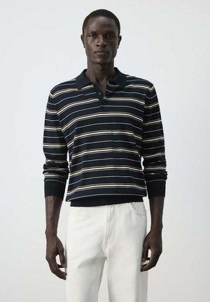 Hombre con una camisa polo negra de manga larga con rayas horizontales blancas y beige y pantalones blancos sobre un fondo liso.