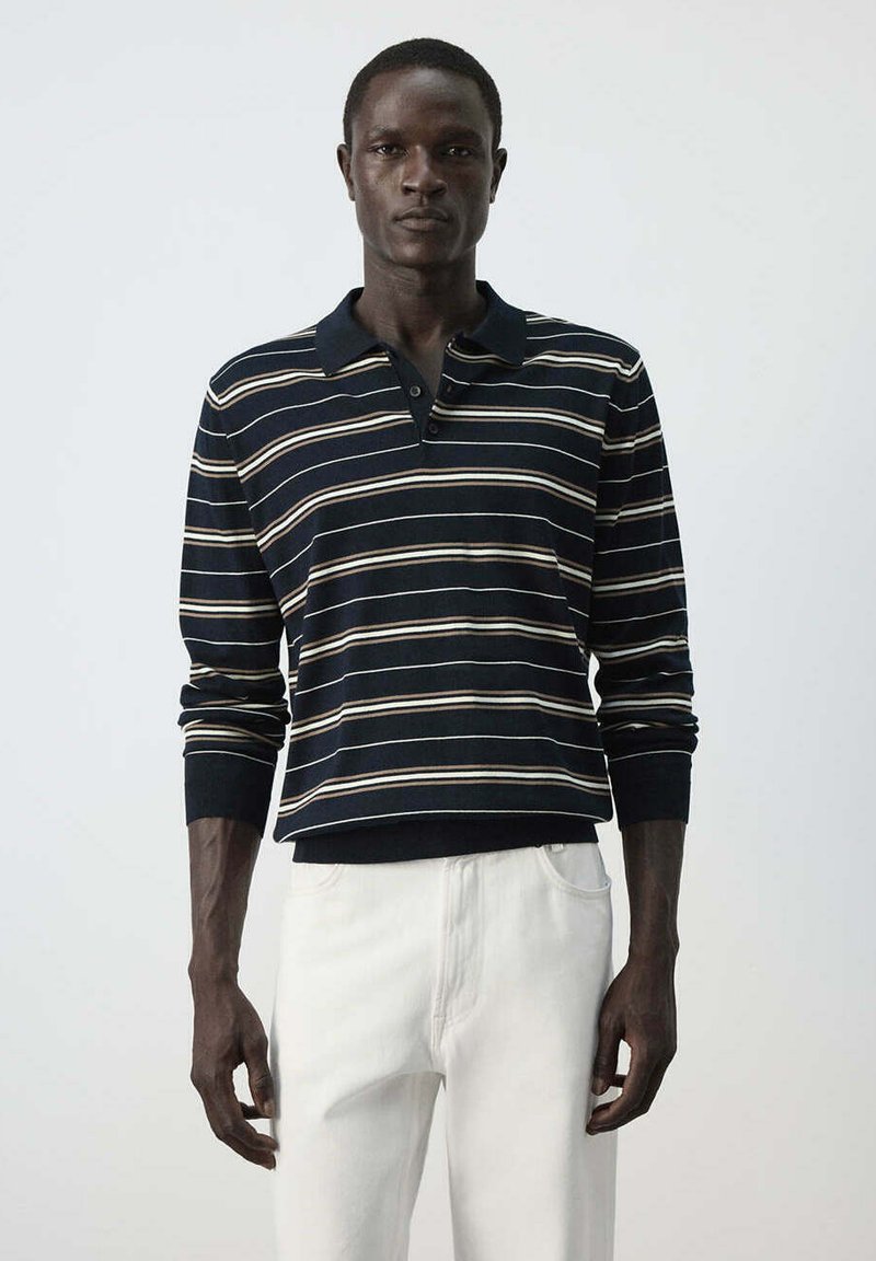 Homme portant un polo noir à manches longues avec des rayures horizontales blanches et beiges et un pantalon blanc sur un fond uni.