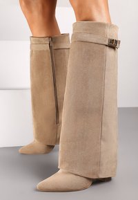 Des bottes en daim beige jusqu'au genou présentent un bout pointu, une fermeture éclair sur le côté et un accent boucle en haut, avec une texture lisse.