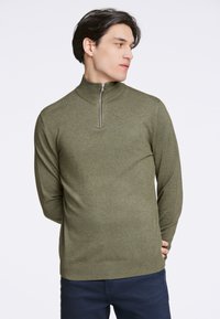 Olivgrüner, langärmeliger Zip-up-Pullover mit hohem Kragen. Gestrickte Textur und figurbetonter Schnitt, mit einem kurzen silbernen Reißverschluss am Hals.