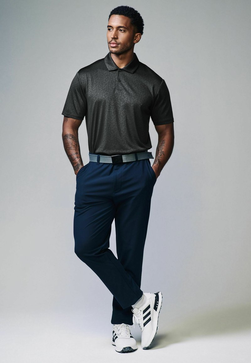 Sort polo skjorte med diskret mønster, mørkeblå bukser, grå bælte, hvide sneakers med sorte akcenter. Model står med hænderne i lommerne.